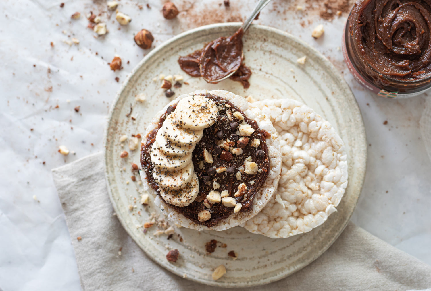 a creative photo of a rice cake topped with the Healthy Alternatives hazelnut spread, an assortment of nuts, and sliced bananas. 